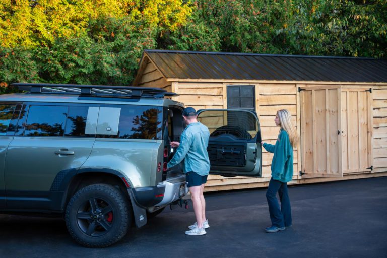 Two people standing next to a suv in front of a shed.