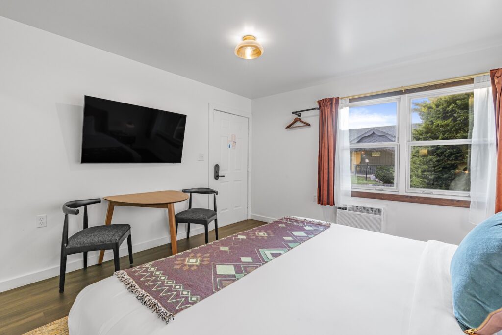 The Superior King room features a plush bed, a mounted TV, and a small table with two chairs. A window adorned with red curtains adds charm, while white walls and wooden flooring create a serene backdrop. A convenient coat hanger completes this welcoming space.