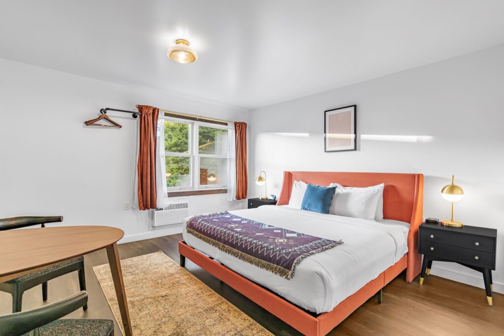 The modern bedroom boasts a Superior King bed with orange upholstery, complemented by white and purple bedding. A round wooden table with chairs adds charm, while a window with curtains and a framed picture complete the serene setting.