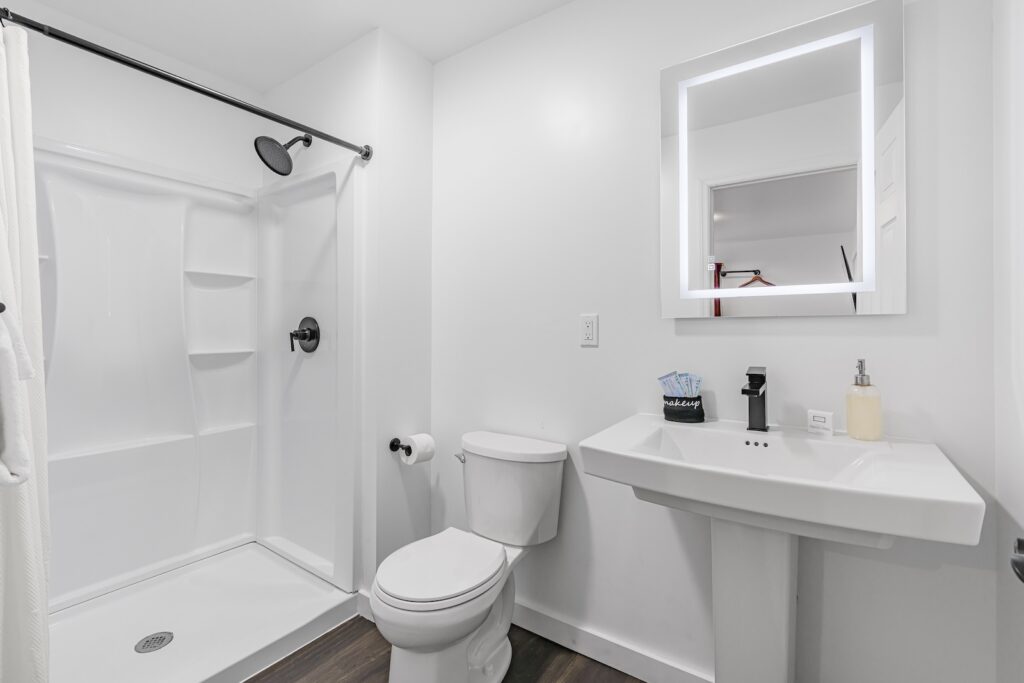 A modern bathroom featuring a shower, toilet, and pedestal sink complements the Superior King's suite. A backlit mirror hangs above the sink, while dark wood-style panels line the floor, enhancing your luxurious retreat experience.