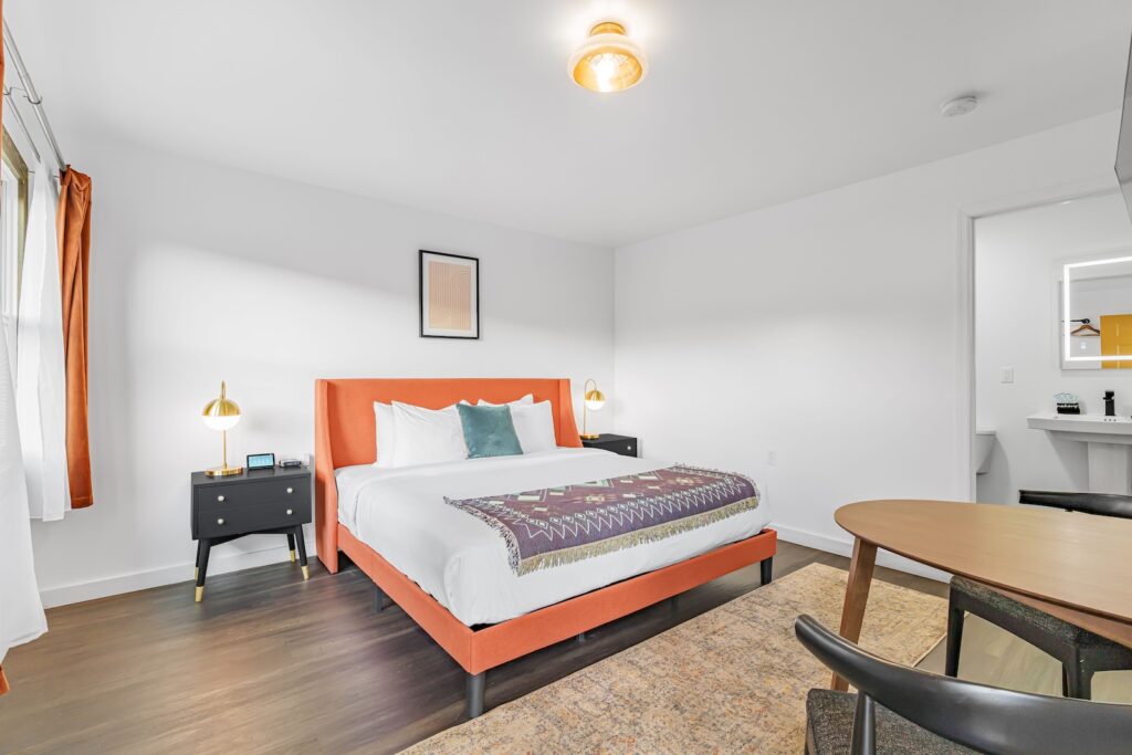 A modern bedroom features a superior orange bed frame with white bedding and a patterned blanket fit for a king. Two black nightstands with lamps flank the bed. In the foreground, a round table and chairs rest on a cozy rug.