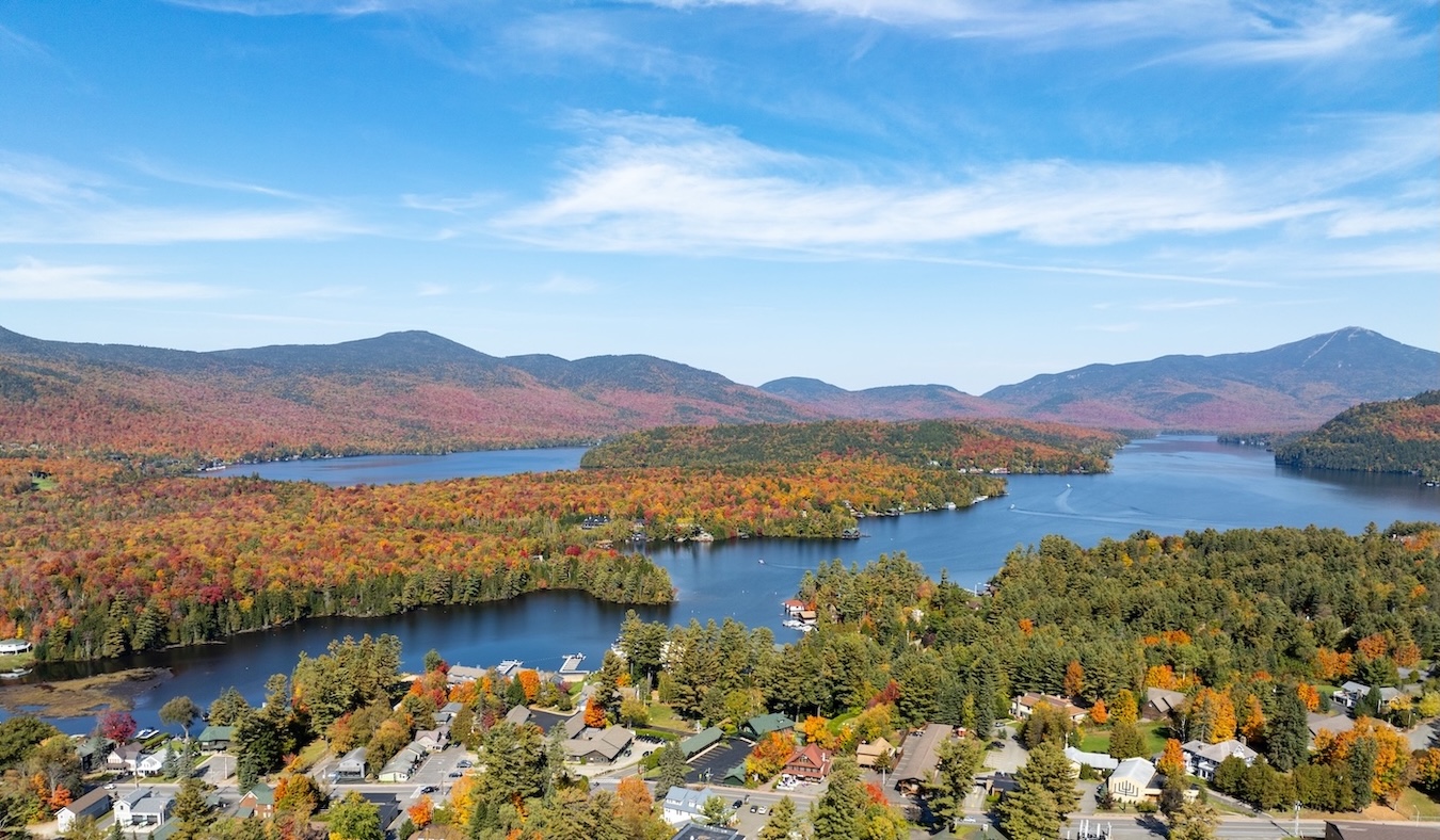 Aerial view of a lake surrounded by forests with autumn foliage and mountains in the background under a blue sky.