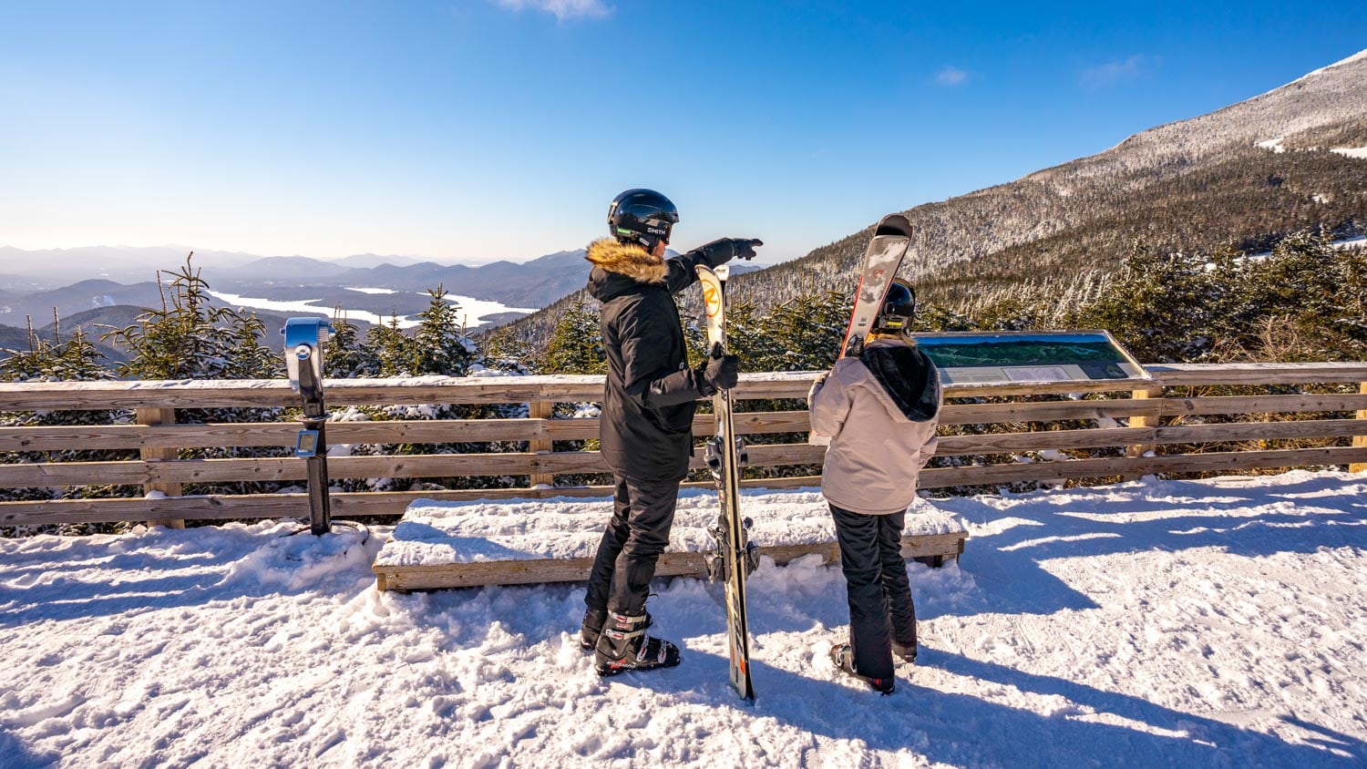 Two people in ski gear stand on a snowy overlook, one holding skis and pointing toward the mountains in the distance under a clear blue sky.